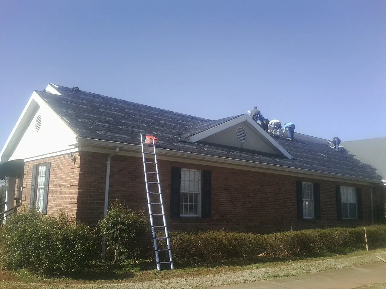 Crew tearing shingles off a brick ranch roof with a ladder set — Metal Roof Repair work by our crew in Interlaken