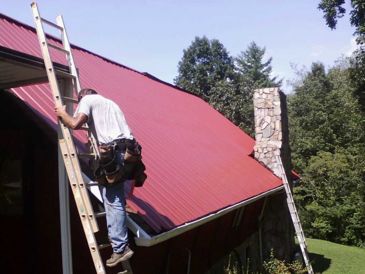 Roofer on a ladder beside a red metal roof and stone chimney — Soffit & Fascia Repair by Roofing Brothers in Interlaken